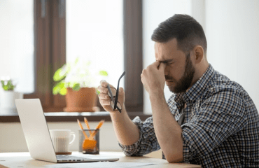 Man sat at desk with laptop, holding the bridge of his nose and holding his glasses in his other hand.